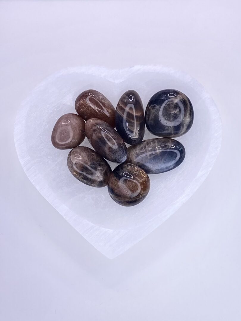 A heart shaped glass bowl with Black Moonstone Tumbled Stones in it.